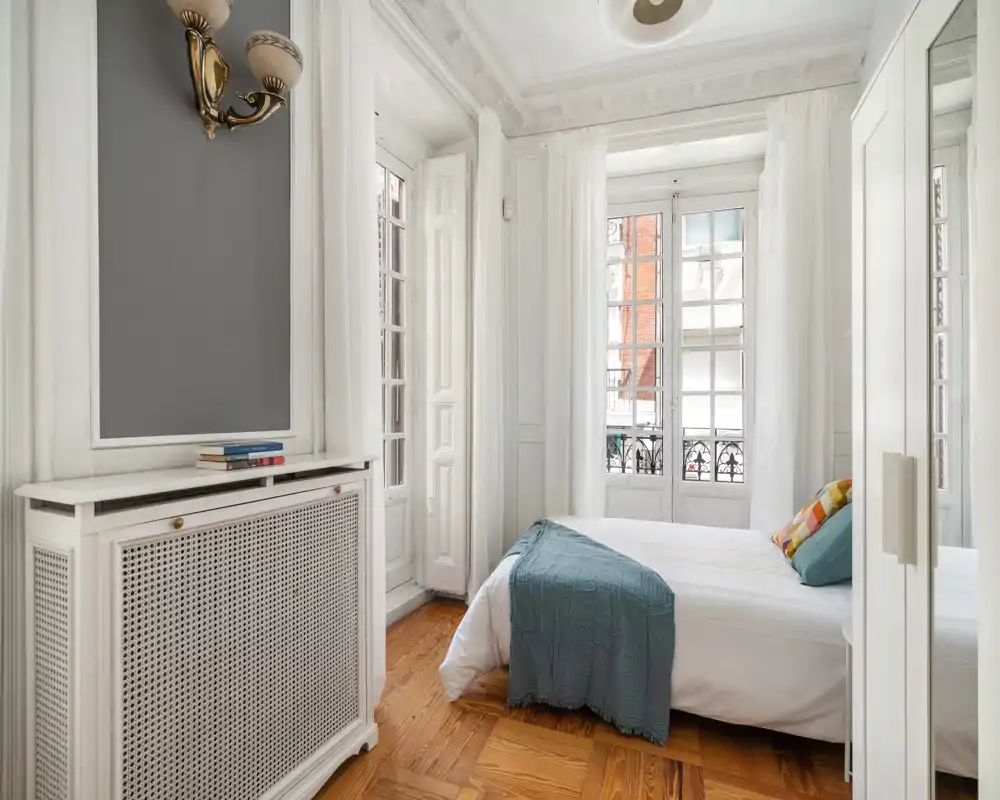 Chambre lumineuse et élégante avec un grand lit bien fait, de hautes fenêtres à voilages, moulures classiques et parquet chaleureux—parfaite pour un séjour confortable en ville.