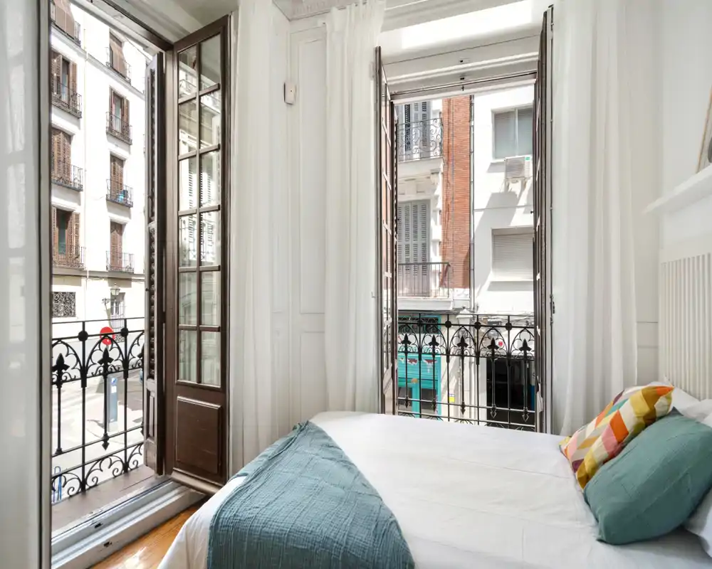 Chambre lumineuse et aérée avec un lit double bien fait, coussins colorés et grandes fenêtres à la française ouvrant sur un balcon urbain.