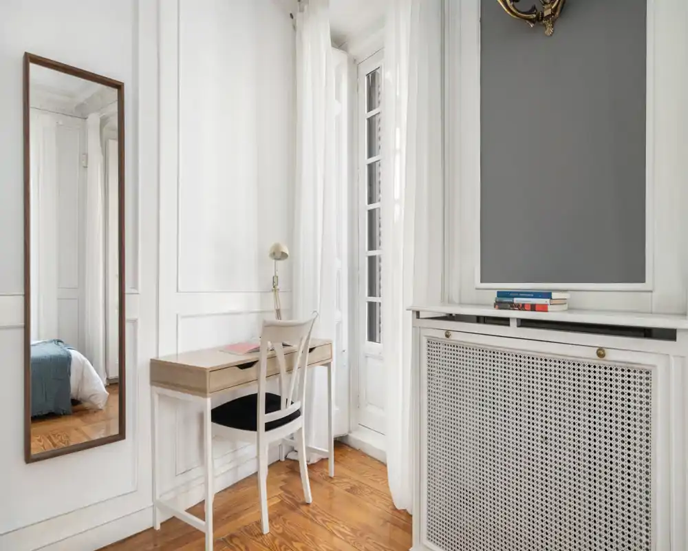 Coin de chambre lumineux et élégant avec un petit bureau, un miroir en pied et un aperçu du lit. Murs blancs, hautes fenêtres et parquet chaleureux offrent une atmosphère aérienne et apaisante.