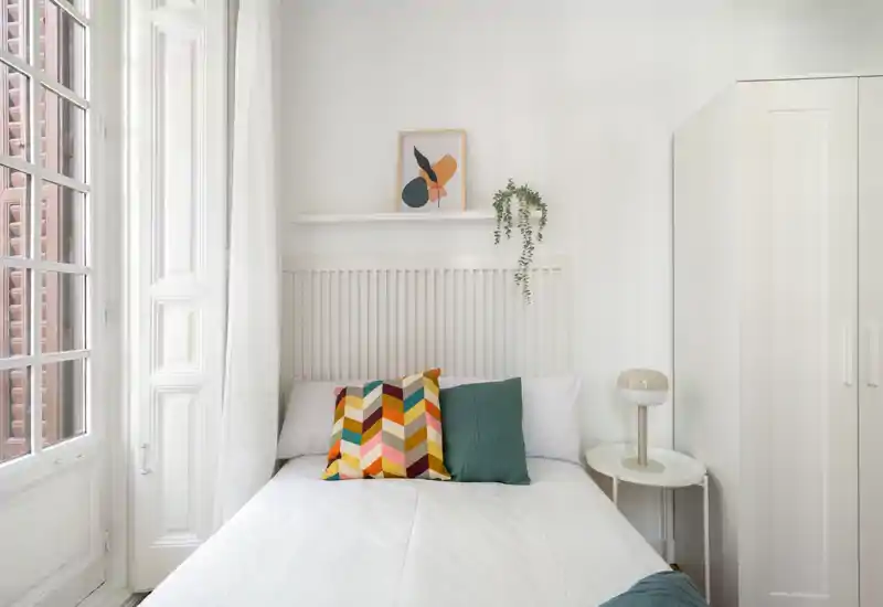 Dormitorio luminoso y con estilo, cama bien hecha con cojines coloridos, mesita con lámpara y armario blanco alto — espacio tranquilo y aireado iluminado por una gran ventana.