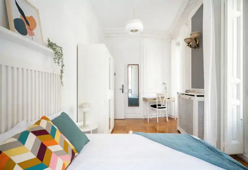 Dormitorio luminoso y con estilo, con cama cómoda, cojines coloridos y una manta verde azulado, mobiliario blanco y un escritorio junto a la ventana — perfecto para una estancia tranquila y moderna.