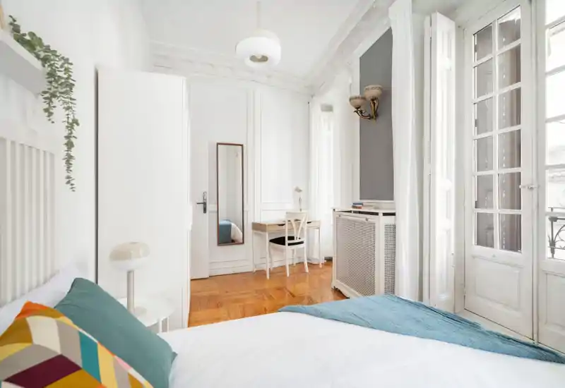 Dormitorio luminoso y elegante con una cama doble cómoda, cojines decorativos y un pequeño escritorio junto a grandes ventanas que aportan abundante luz natural.