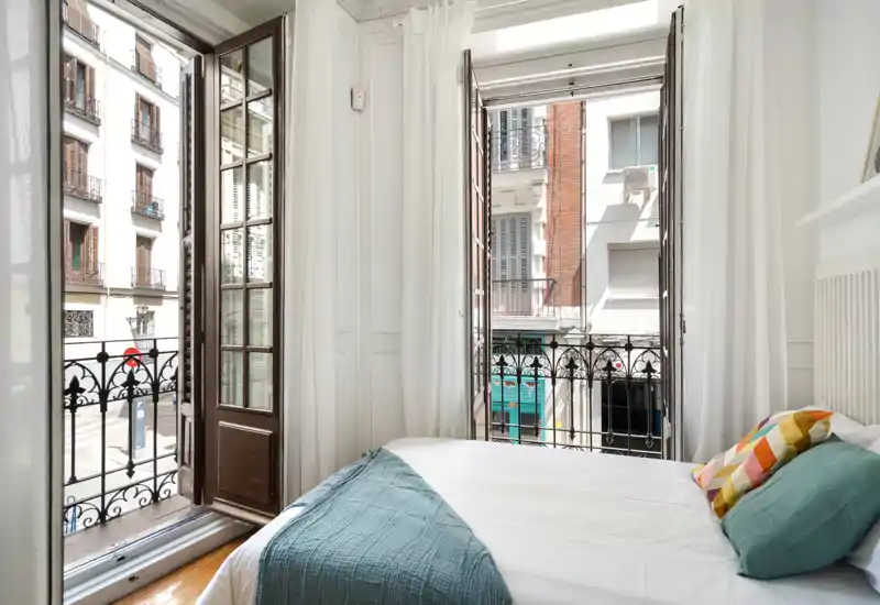 Dormitorio luminoso y aireado con una cama doble bien hecha, cojines de colores y grandes ventanas francesas que dan a un balcón urbano.