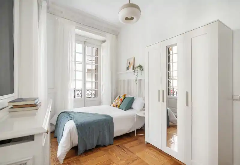Dormitorio luminoso y aireado con cama doble, cojines decorativos y una manta turquesa. Grandes ventanas inundan la estancia de luz natural y un armario blanco con espejo amplía la sensación de espacio.