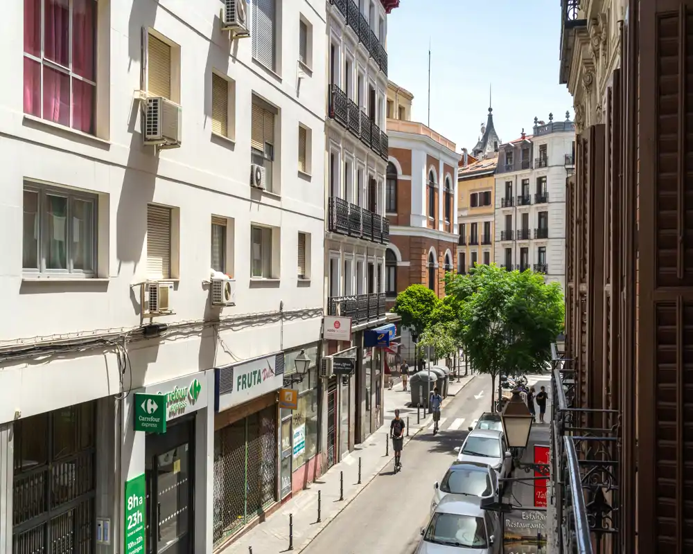 Calle urbana soleada vista desde un balcón: vía estrecha, coches aparcados, pequeños comercios y peatones con luz diurna — ideal para mostrar la ubicación y el entorno.