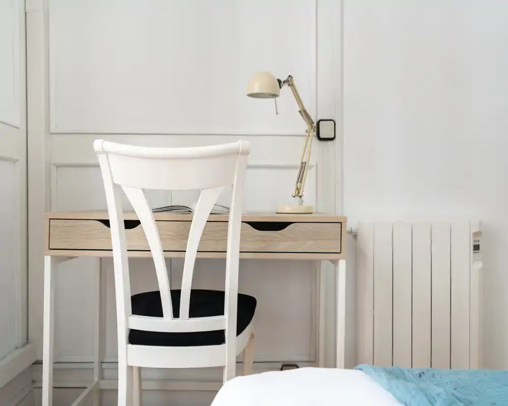 Rincón de dormitorio luminoso y minimalista con un pequeño escritorio de madera, silla blanca y lámpara; se aprecia una porción de la cama con una manta azul claro en primer plano.