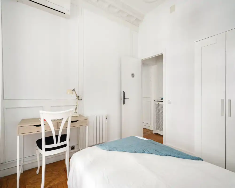 Dormitorio luminoso y minimalista con una cama cómoda, un pequeño escritorio y un armario. Los techos altos y las molduras decorativas añaden un aire refinado.