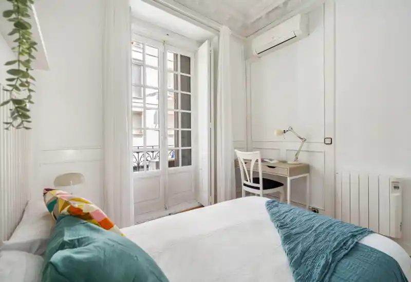 Dormitorio luminoso y despejado con cama doble cómoda, cojines decorativos y una manta texturizada. Gran ventana francesa que aporta mucha luz natural y un pequeño escritorio para un rincón de trabajo acogedor.