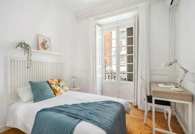 Dormitorio luminoso y elegante con cama doble vestida en blanco, cojines de colores y una manta turquesa. Gran ventana estilo francés que aporta luz natural; escritorio pequeño y mesita de noche completan la estancia.