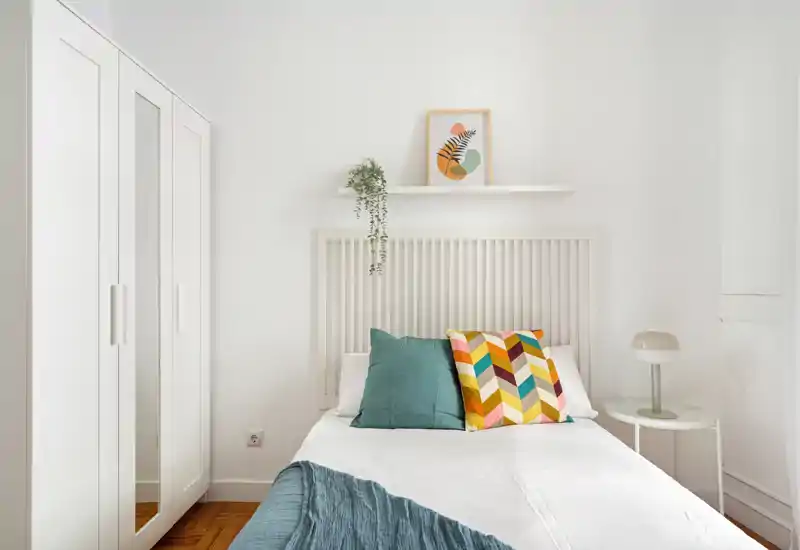 Dormitorio luminoso y minimalista con la cama bien hecha, cojines decorativos coloridos y una pequeña lámpara de mesilla. Paredes blancas y armario crean una sensación limpia y aireada, perfecta para anuncios.