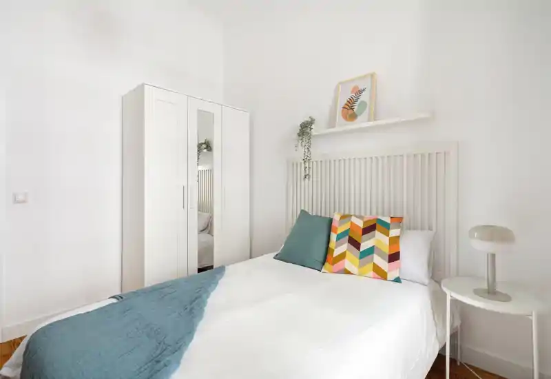 Dormitorio luminoso y minimalista con cabecero blanco de lamas, cojines de colores y mesita con lámpara. Estilo limpio, armario con puerta espejo y suave luz natural, ideal para anuncios.