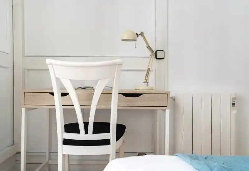 Angolo di camera luminoso e minimale con scrivania in legno, sedia e termosifone; una piccola porzione del letto e una coperta azzurra sono visibili in primo piano.