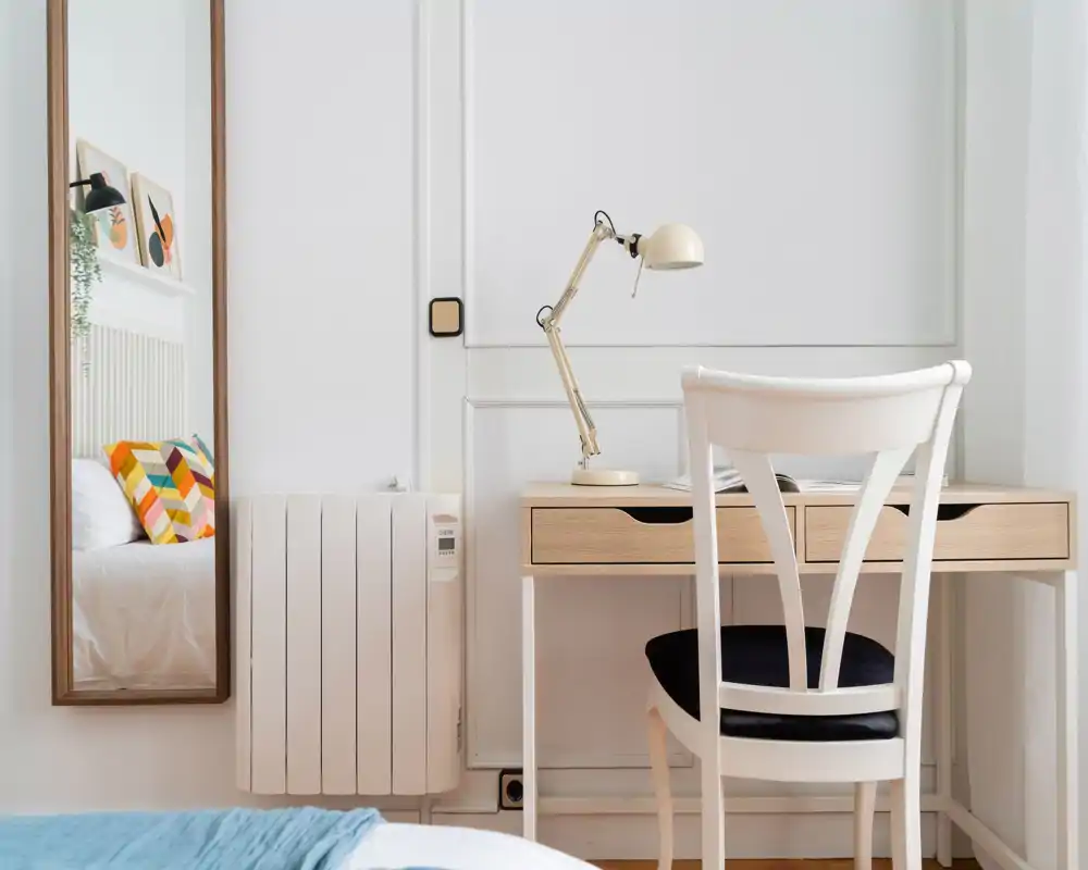 Angolo camera luminoso e ben curato con letto visibile (in parte riflesso in uno specchio alto), una scrivania in legno e una sedia con lampada da tavolo—arredo minimal e stile nordico.