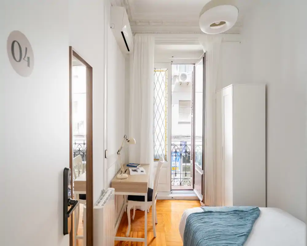 Camera da letto luminosa e compatta con letto comodo, pavimento in parquet, una scrivania vicino alla finestra e accesso a un balconcino — ideale per viaggiatori singoli o smart working.