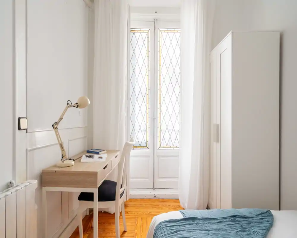 Camera da letto luminosa e minimal con letto ordinato, scrivania in legno e sedia vicino a una finestra alta che assicura molta luce naturale.