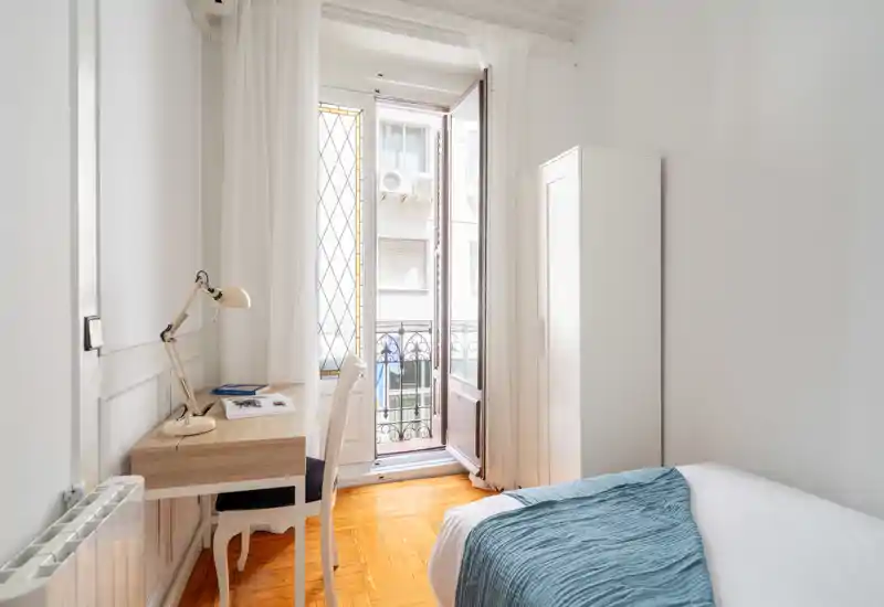 Dormitorio luminoso y minimalista con cama cómoda, suelo de parquet y un escritorio junto a una gran ventana/puerta que da a un pequeño balcón. Paredes blancas y luz natural crean un ambiente aireado y tranquilo, ideal para alquileres de corta estancia.