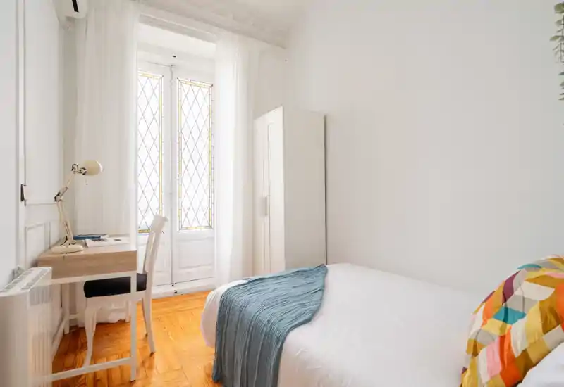 Dormitorio luminoso y minimalista con una cama cómoda, textiles suaves y un pequeño escritorio junto a la ventana que deja entrar mucha luz natural.