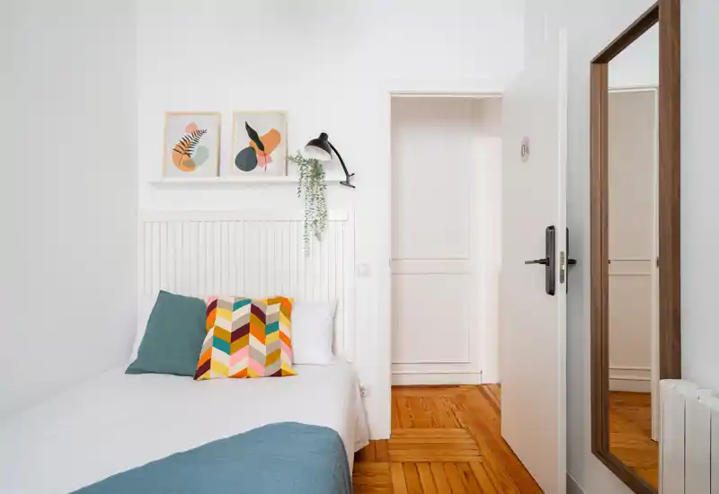 Dormitorio luminoso y moderno con cama bien hecha, cojines decorativos coloridos y arte de pared minimalista. Suelo de madera cálida y espejo de cuerpo entero junto a la puerta.