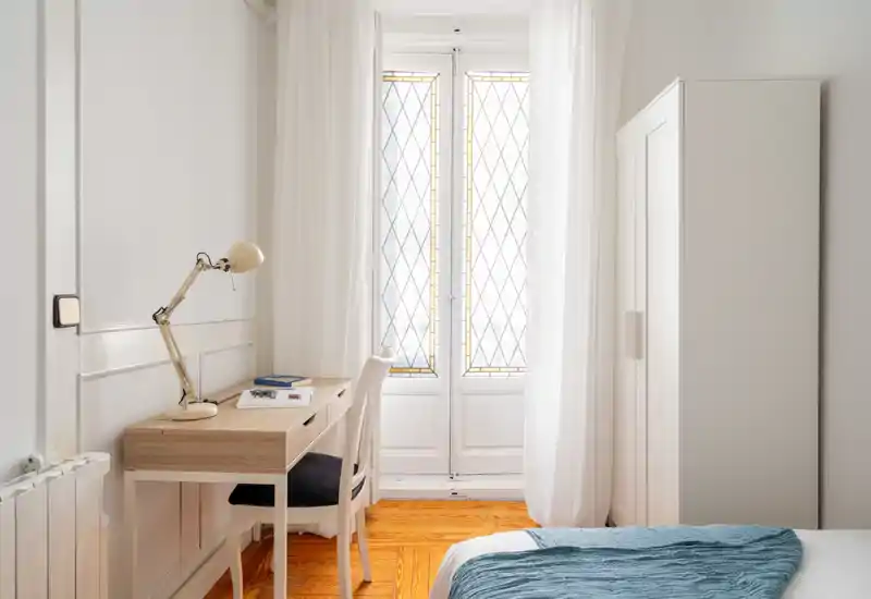 Angolo camera da letto luminoso e minimale con letto ordinato, armadio bianco e una piccola scrivania in legno vicino a porte-finestre con vetri decorati che lasciano entrare molta luce naturale.