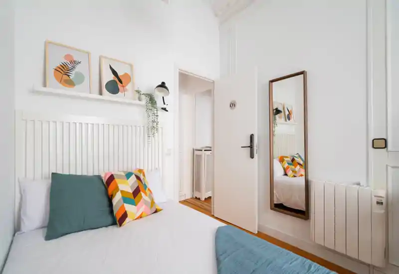 Dormitorio luminoso y con estilo, cama bien hecha, cojines coloridos y arte de pared minimalista. Paredes blancas y luz natural que crean un ambiente fresco y acogedor.