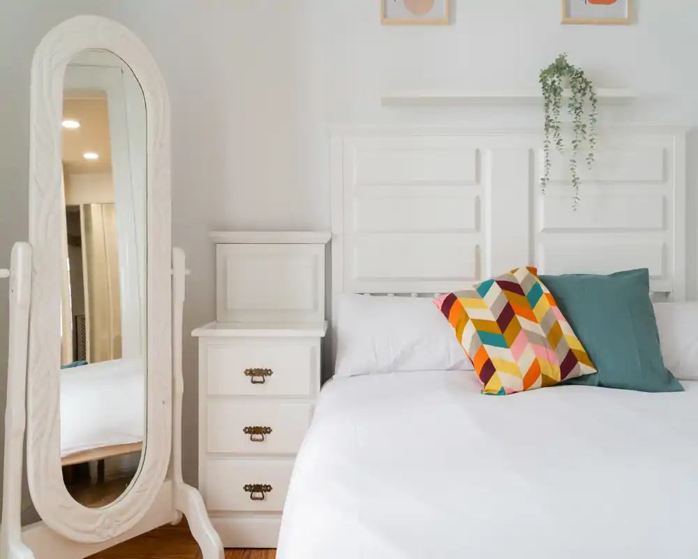 Camera da letto luminosa e ordinata con testiera bianca, lenzuola impeccabili e cuscini colorati; uno specchio a figura intera decorativo e un comodino completano l'ambiente moderno.