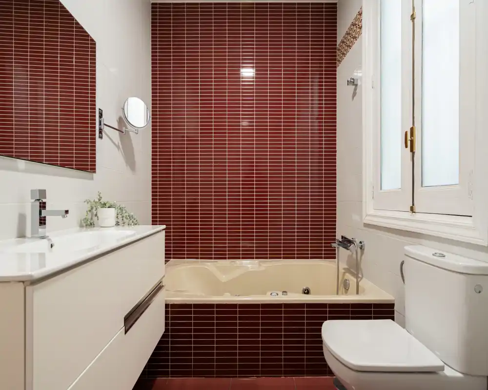 Bagno moderno con vasca incassata davanti a una parete di piastrelle rosso scuro, mobile lavabo bianco con specchio e wc accanto a una finestra opaca. Stile pulito e contemporaneo con contrasto delle piastrelle.