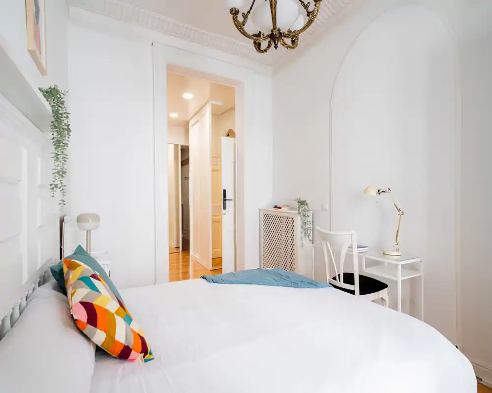 Camera da letto luminosa ed elegante con un grande letto bianco, cuscino colorato e una piccola scrivania: perfetta per soggiorni riposanti.