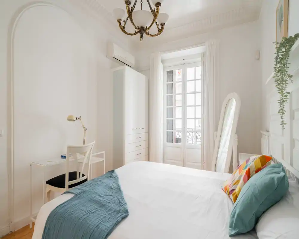 Camera da letto luminosa ed elegante con letto bianco, cuscini decorativi e finestrone che lascia entrare molta luce naturale; arredamento pulito e minimale con armadio, scrivania e specchio a tutta altezza.