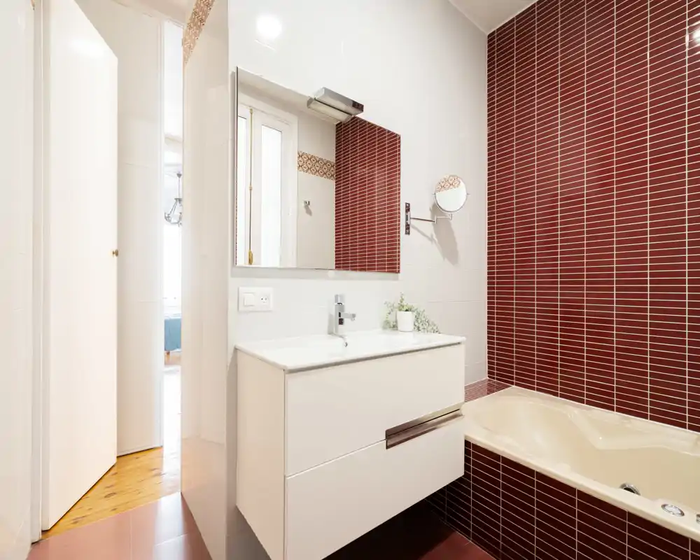 Bagno moderno e luminoso con mobile lavabo bianco, specchio e vasca ad angolo incorniciata da piastrelle verticali rosso scuro. Linee pulite e illuminazione curata rendono l'ambiente fresco e ben tenuto.