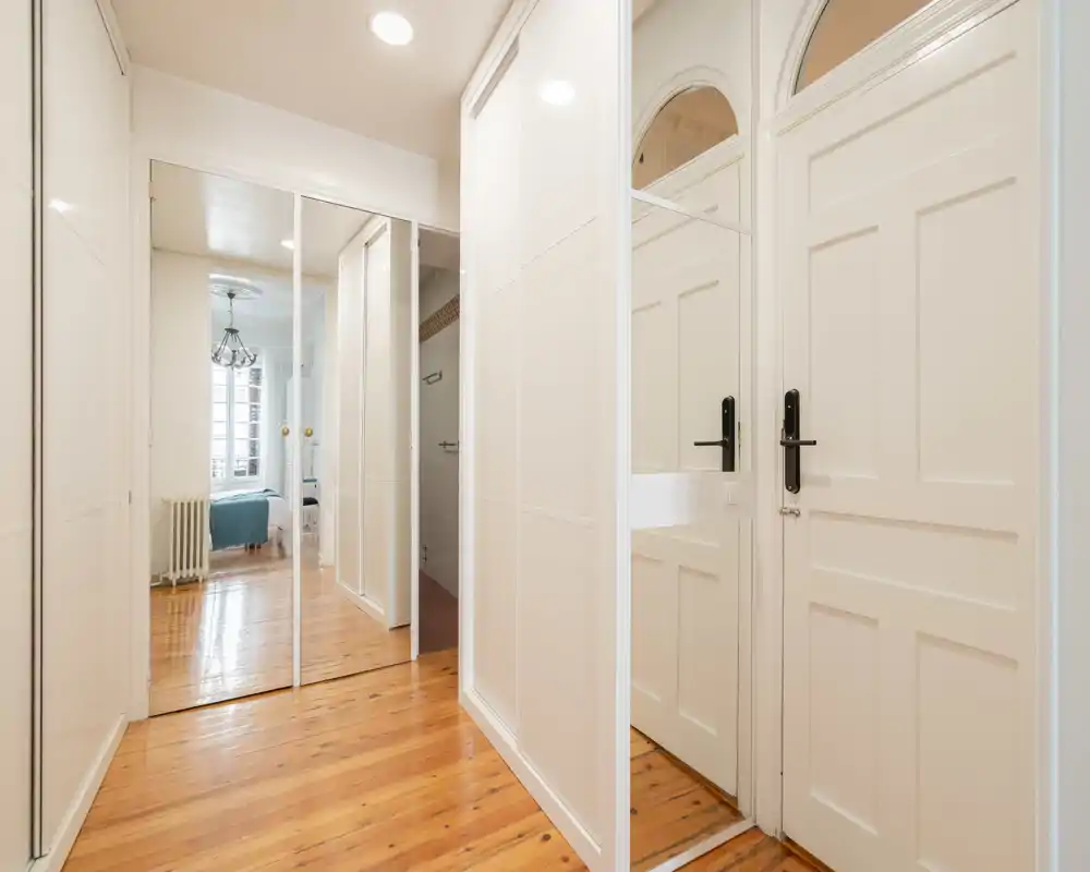 Corridoio luminoso che conduce a una camera da letto visibile attraverso una porta aperta, con armadi a specchio e pavimento in legno; ambiente pulito e arioso adatto a un annuncio.