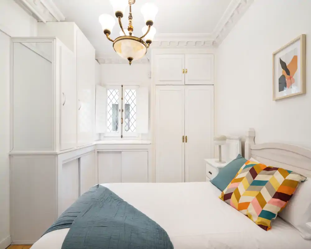 Dormitorio luminoso y elegante con armario empotrado blanco, molduras decorativas y una lámpara de techo clásica. La cama está bien hecha con cojines coloridos y una manta verde azulado que aporta calidez.