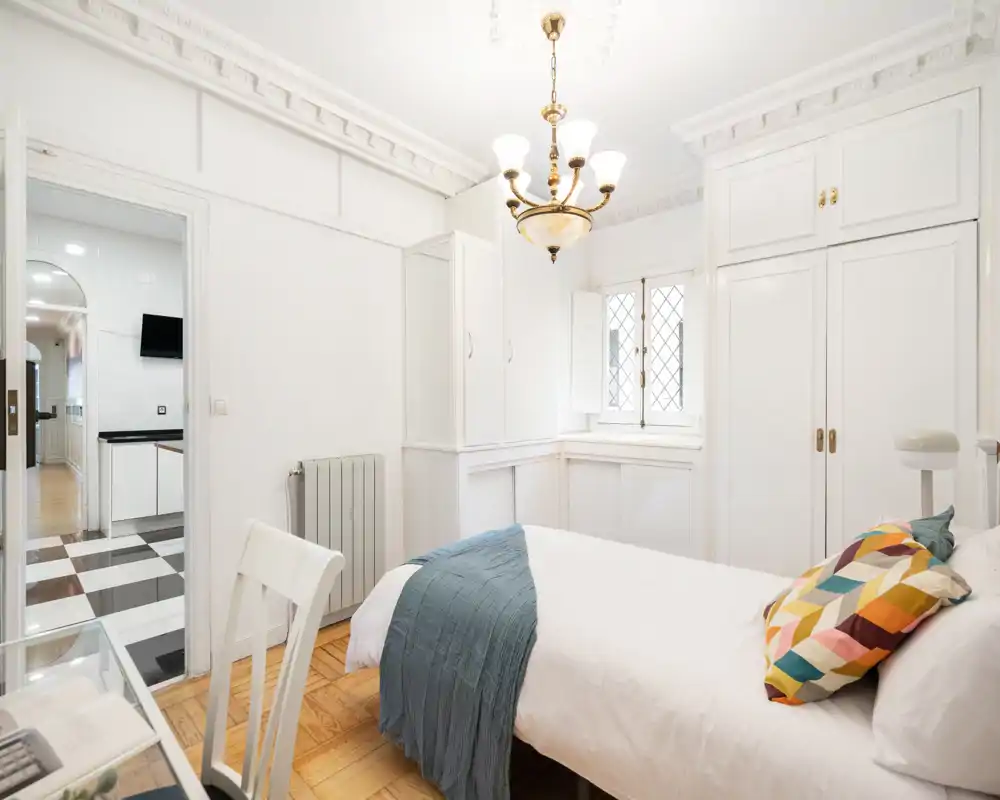 Dormitorio luminoso y elegante con cama doble cómoda, cojines decorativos y armarios empotrados blancos; la luz natural y los detalles del techo crean un ambiente acogedor y refinado.