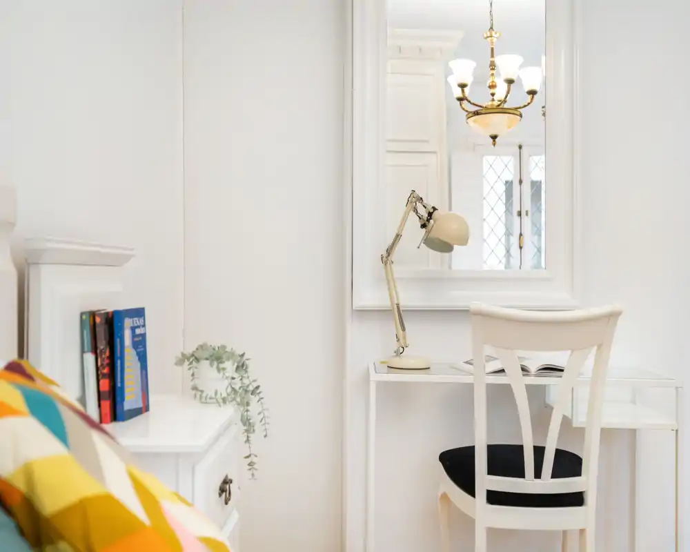 Rincón de dormitorio luminoso y minimalista con un pequeño escritorio blanco, silla y mesita. La imagen muestra una almohada decorativa y parte de la cama, una lámpara de escritorio y un espejo que refleja detalles del techo.