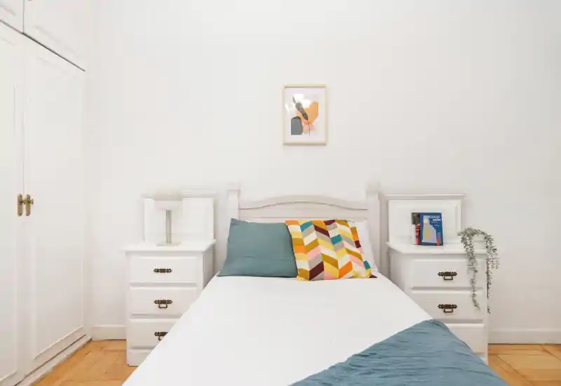 Camera da letto luminosa e minimal con un letto singolo ben rifatto affiancato da comodini bianchi coordinati, cuscini colorati e luce naturale soffusa.
