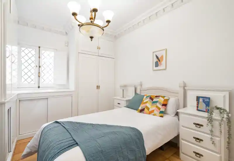Dormitorio individual luminoso y acogedor con cama bien hecha, cojines coloridos y armarios blancos clásicos. La luz natural entra por la ventana con contraventanas y las molduras y la lámpara aportan carácter.