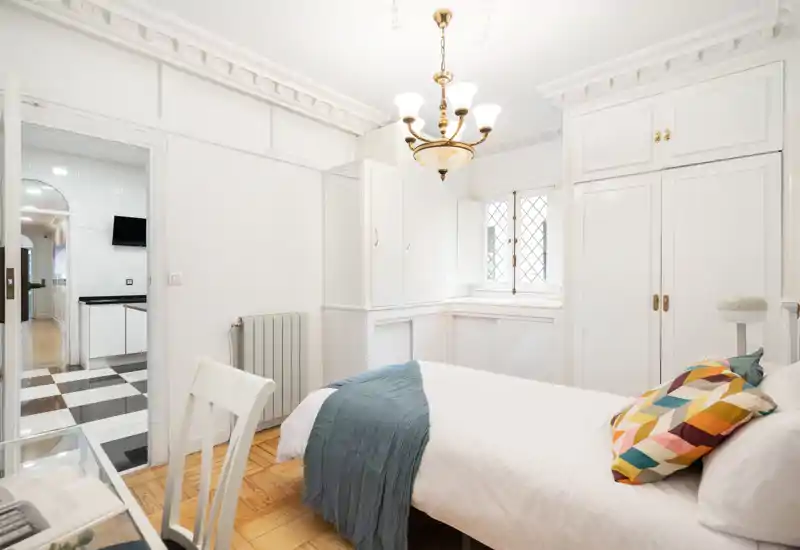 Dormitorio luminoso y elegante con cama doble cómoda, cojines decorativos y armarios empotrados blancos; la luz natural y los detalles del techo crean un ambiente acogedor y refinado.