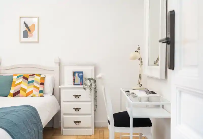 Dormitorio luminoso y aireado con la cama bien hecha, cojines coloridos de estampado geométrico, una mesita blanca y un pequeño escritorio con lámpara — un espacio tranquilo y bien decorado, ideal para estancias cortas.