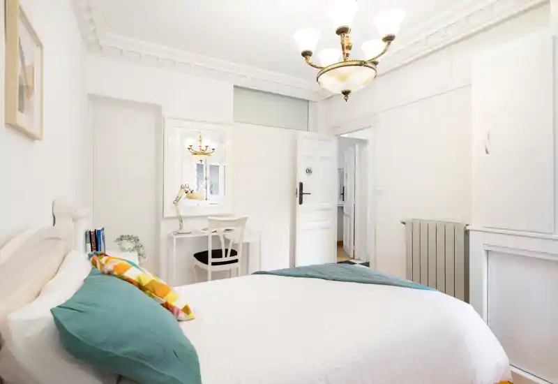 Dormitorio luminoso y bien decorado con una cama blanca amplia, cojines coloridos y un pequeño escritorio junto a la pared. Molduras clásicas y una lámpara de techo crean un ambiente atractivo y acogedor.