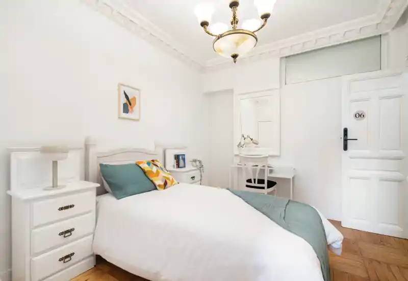 Dormitorio individual luminoso y elegante con mobiliario blanco, cojines decorativos y un pequeño escritorio — ideal para una estancia cómoda.