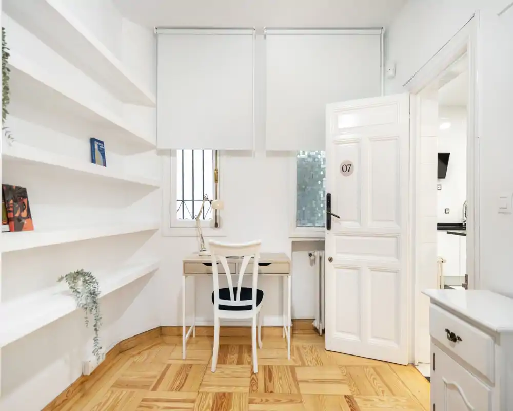 Spazio living luminoso e minimale con scaffalature integrate e una piccola zona studio — ideale per leggere o lavorare. Pavimento in legno chiaro, pareti bianche e una sedia ordinata vicino alla finestra.