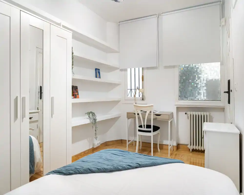 Camera da letto luminosa e ben arredata con letto rifinito in primo piano, scaffalature integrate, armadio con specchio e una piccola scrivania vicino alla finestra — ideale per una persona o una coppia.