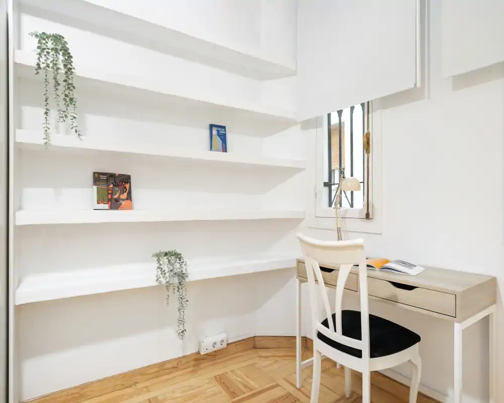 Angolo luminoso soggiorno/studio con scaffalature bianche incassate, una scrivania compatta in legno e una sedia vicino alla finestra; arredo minimal con piccole piante e pavimento in parquet.