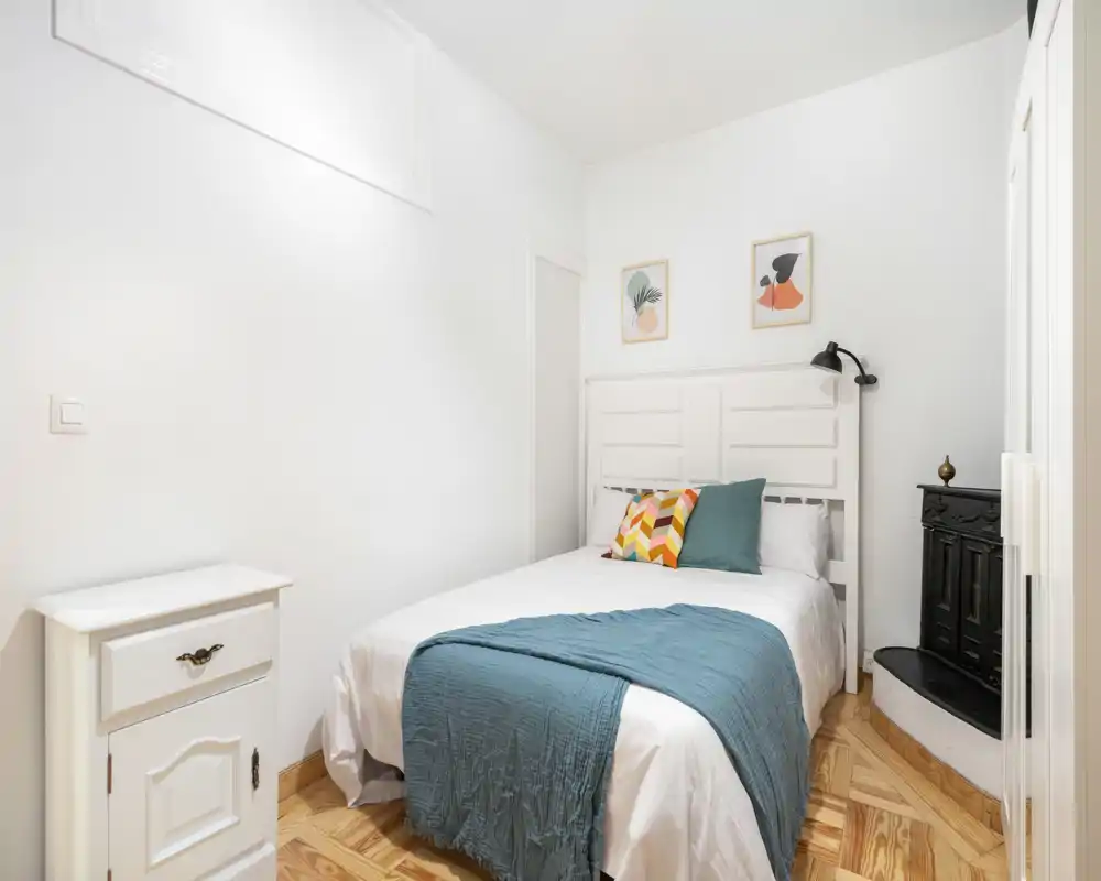 Camera da letto luminosa e compatta con testata bianca a pannelli, cuscini decorativi e un plaid turchese su un letto matrimoniale ben rifatto. Pavimento in parquet e arredo minimal creano un ambiente accogliente.