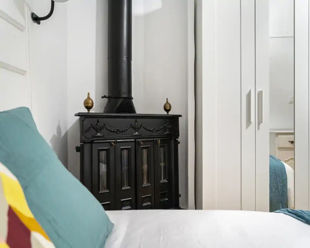 Angolo camera da letto luminoso e accogliente con un letto comodo e cuscini colorati, affiancato da una decorativa stufa in ghisa nera e armadi bianchi.
