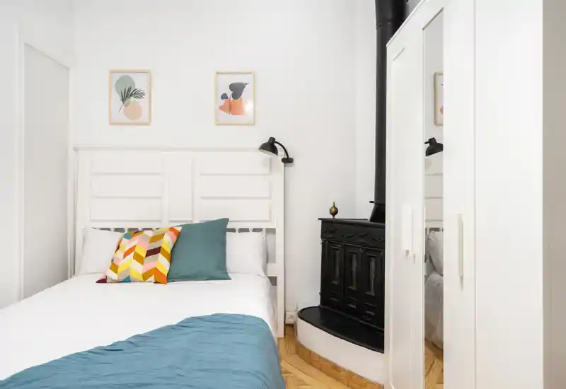 Dormitorio luminoso y bien preparado con cabecero blanco, cojines coloridos y una manta turquesa. Un armario con espejo y una estufa de hierro decorativa en un lateral aportan carácter.