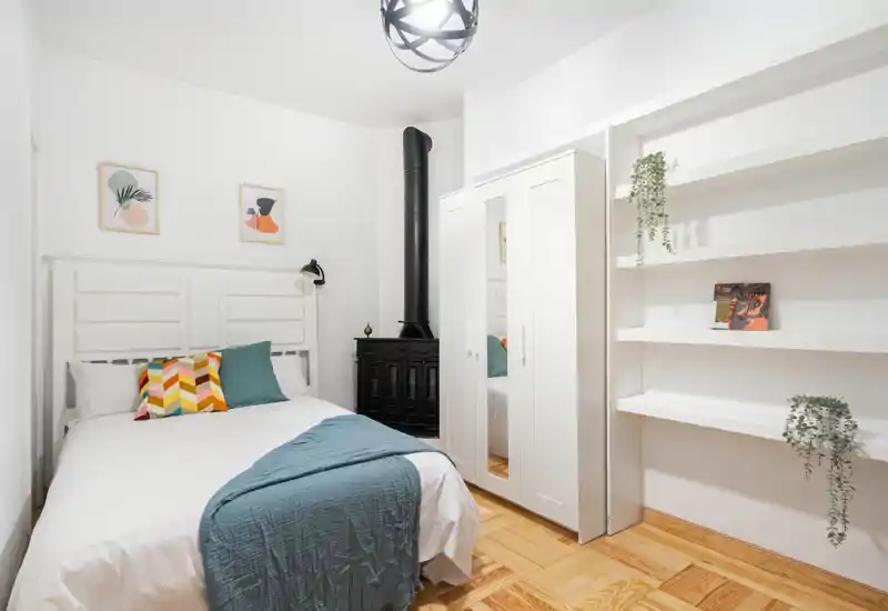 Dormitorio luminoso y estiloso con cama doble, cojines estampados, plaid azul y armario blanco. Suelo de parquet y estanterías integradas aportan calidez y funcionalidad.