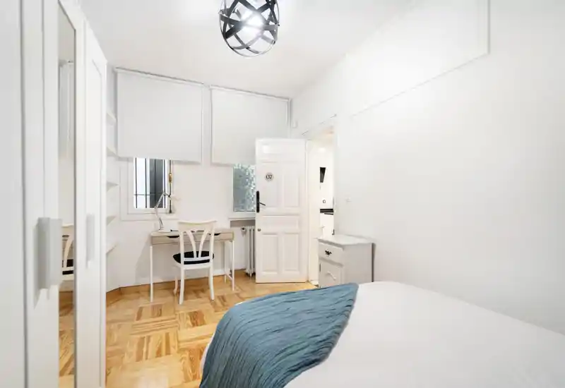 Dormitorio luminoso y minimalista con suelo de parqué, cama hecha con una manta azul y un pequeño escritorio junto a la ventana — limpio, moderno y bien iluminado.