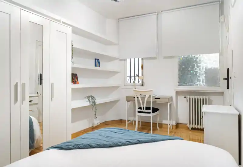 Dormitorio luminoso y bien decorado con la cama arreglada en primer plano, estanterías empotradas, armario con espejo y un pequeño escritorio junto a la ventana — ideal para una persona o pareja.