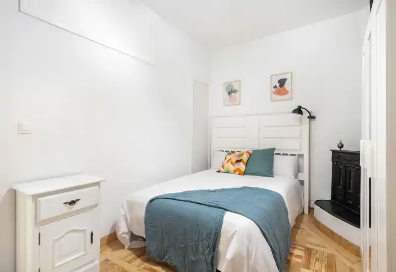 Dormitorio luminoso y compacto con cabecero blanco de paneles, cojines estampados y una manta verde azulado sobre una cama doble ordenada. Suelo de parquet y decoración minimalista que aportan calidez.