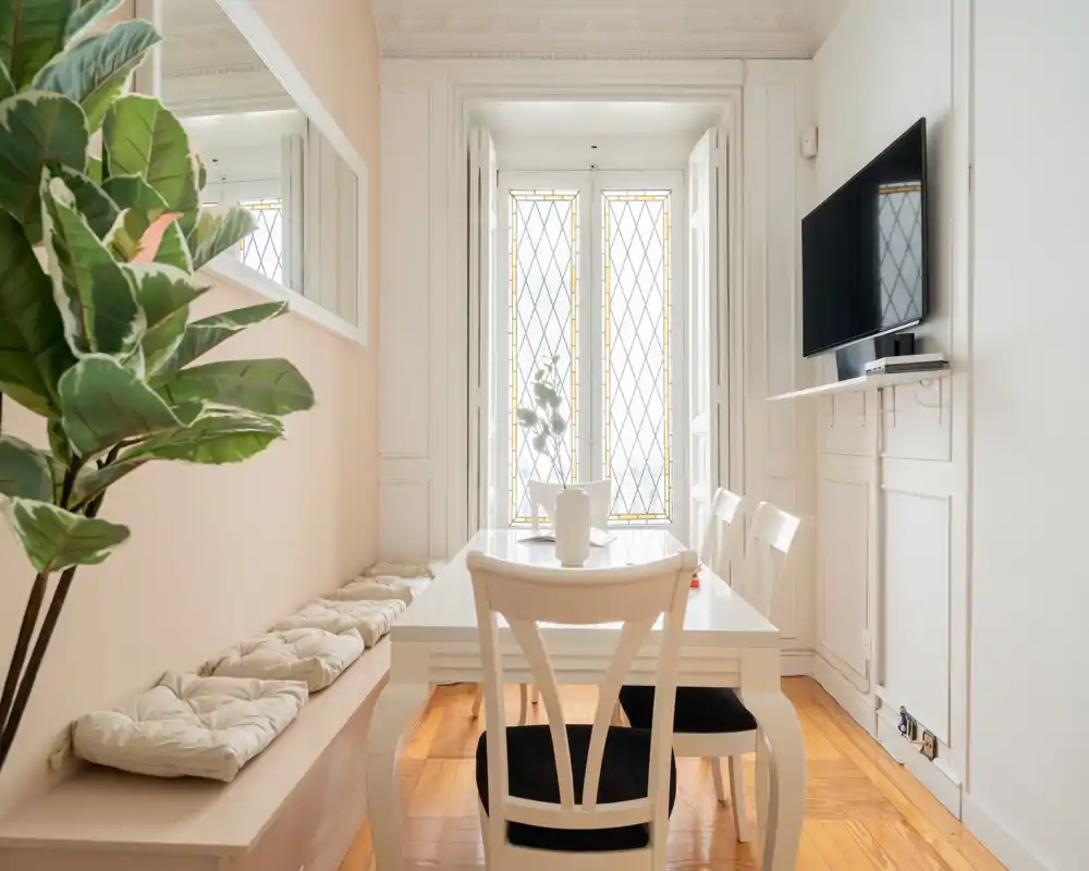Agradable rincón de comedor luminoso con mesa blanca, banco acolchado y sillas frente a una gran ventana decorativa; una planta añade frescura.